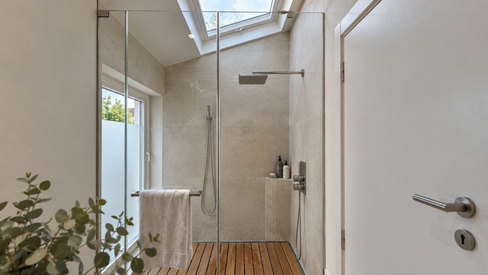 Douche du projet à Colombes, éclairée par une fenêtre de toit, avec paroi en verre et revêtement beige.