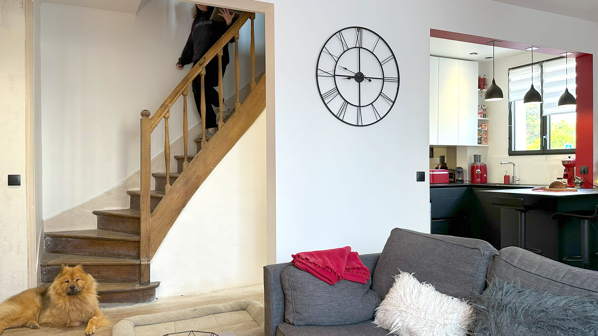Intérieur moderne avec escalier en bois, grand horloge murale, salon et cuisine rouge.